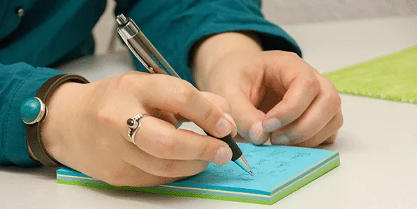 Eine Frau in einem grünen Blazer erstelllt Notizen auf einem Post it. Sie hält einen Kugelschreiber.