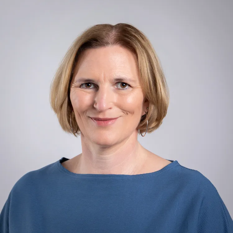 Portrait von Julia Varga vor einem neutralen Hintergrund. Sie trägt einen blauen Pullover und lächelt in die Kamera.